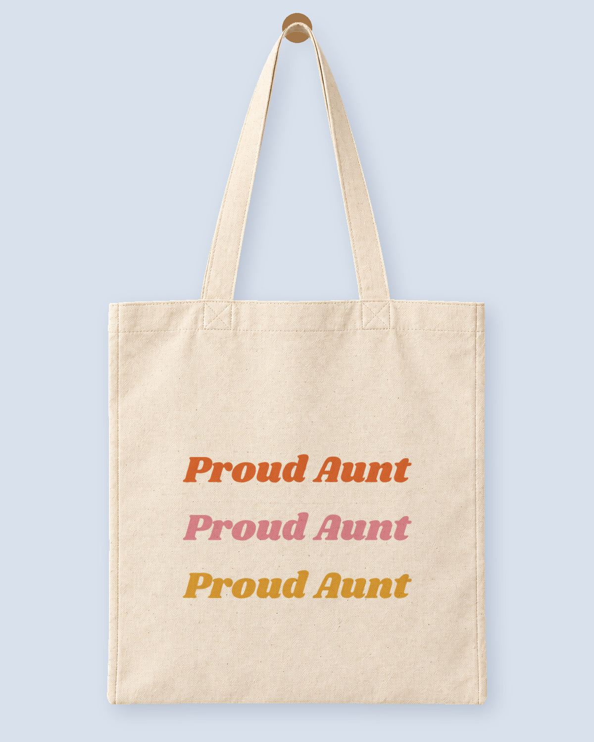 Natural canvas tote printed with “Proud Aunt”, hanging on a wooden peg.