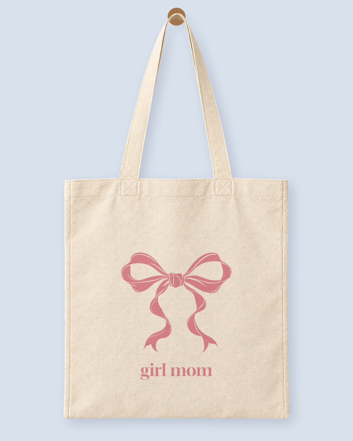 Natural canvas tote printed with “girl mom”, hanging on a wooden peg.
