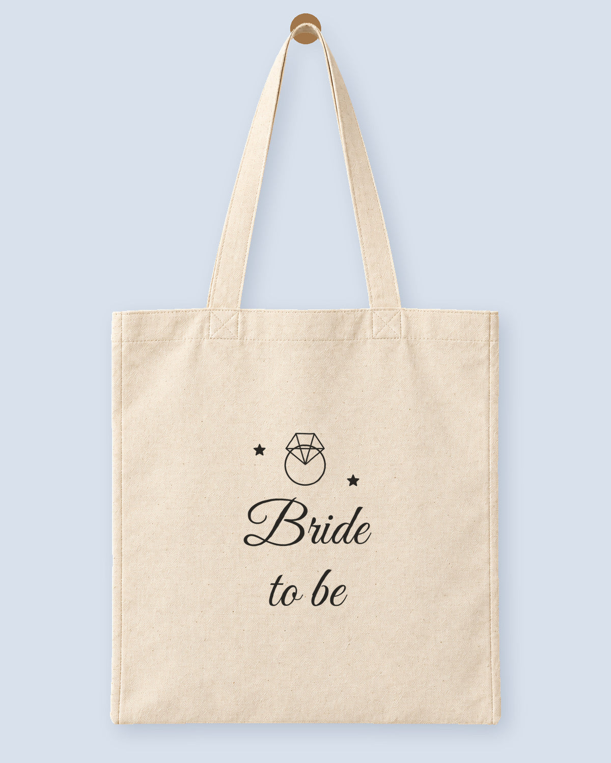Natural canvas tote printed with “Bride to be”, hanging on a wooden peg.