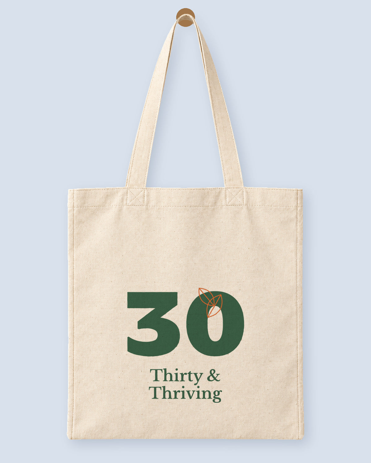 Natural canvas tote printed with “Thirty & Thriving”, hanging on a wooden peg.