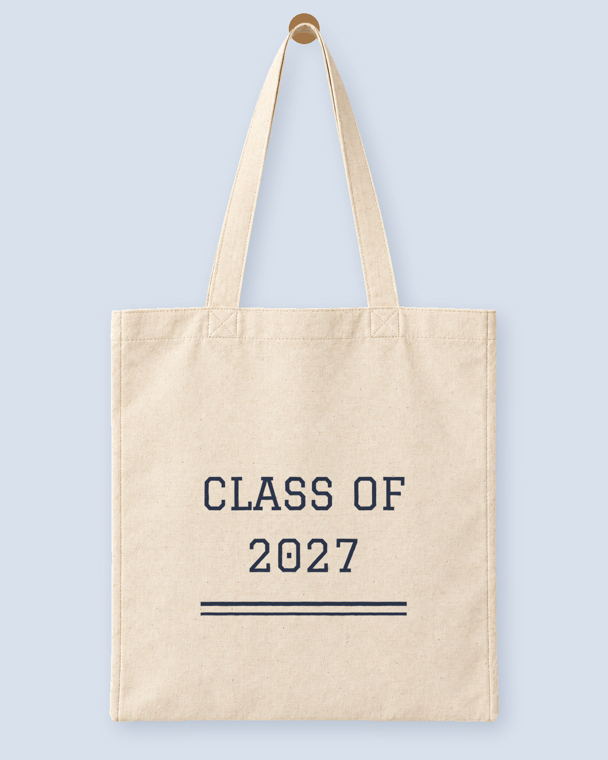Natural canvas tote printed with “CLASS OF 2027”, hanging on a wooden peg.