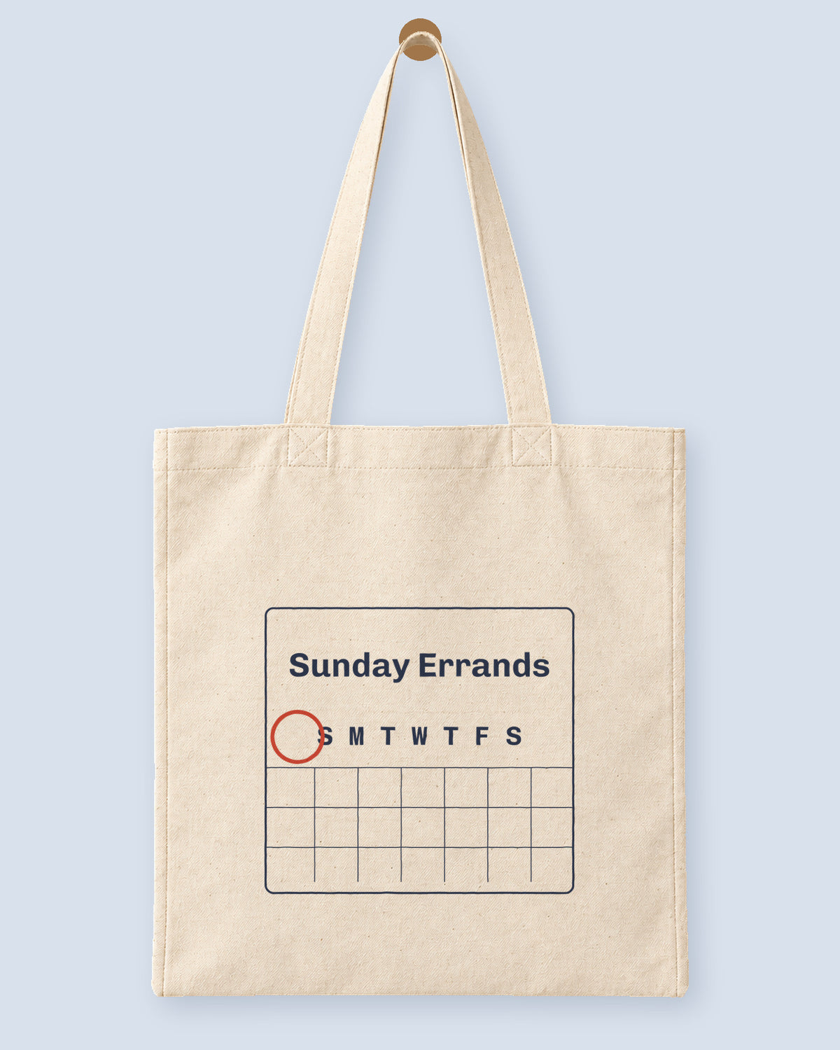 Natural canvas tote printed with “Sunday Errands”, hanging on a wooden peg.