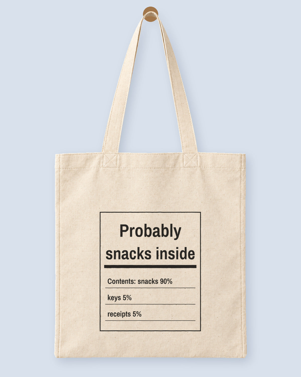 Natural canvas tote printed with “Probably snacks inside”, hanging on a wooden peg.