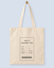 Natural canvas tote printed with “DAD'S GROCERY RUN”, hanging on a wooden peg.