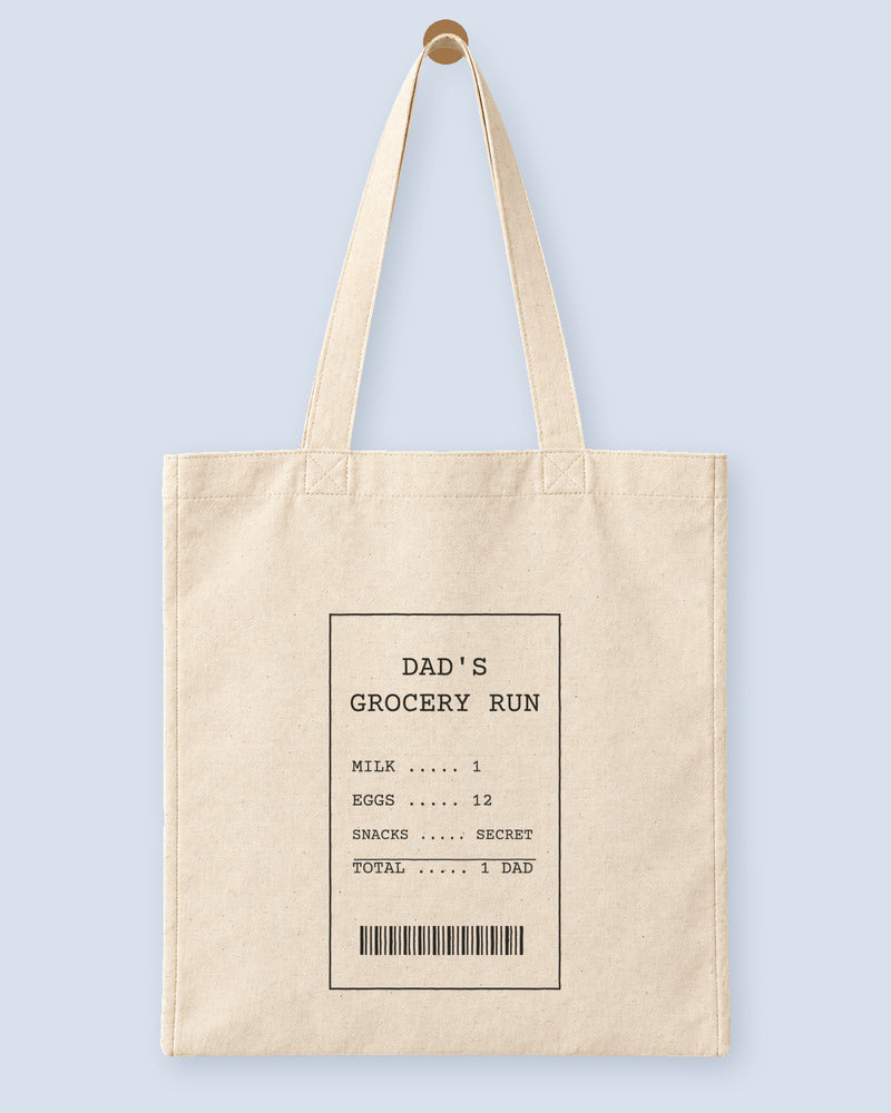 Natural canvas tote printed with “DAD'S GROCERY RUN”, hanging on a wooden peg.