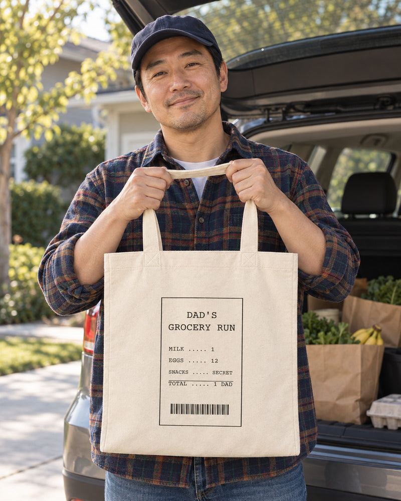 East asian man carrying a natural canvas tote printed with “DAD'S GROCERY RUN” in black. Styled digital image.