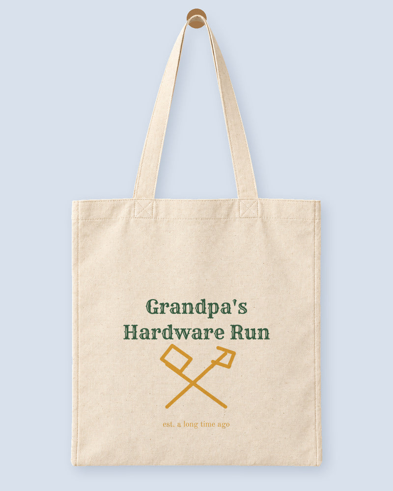 Natural canvas tote printed with “Grandpa's Hardware Run”, hanging on a wooden peg.