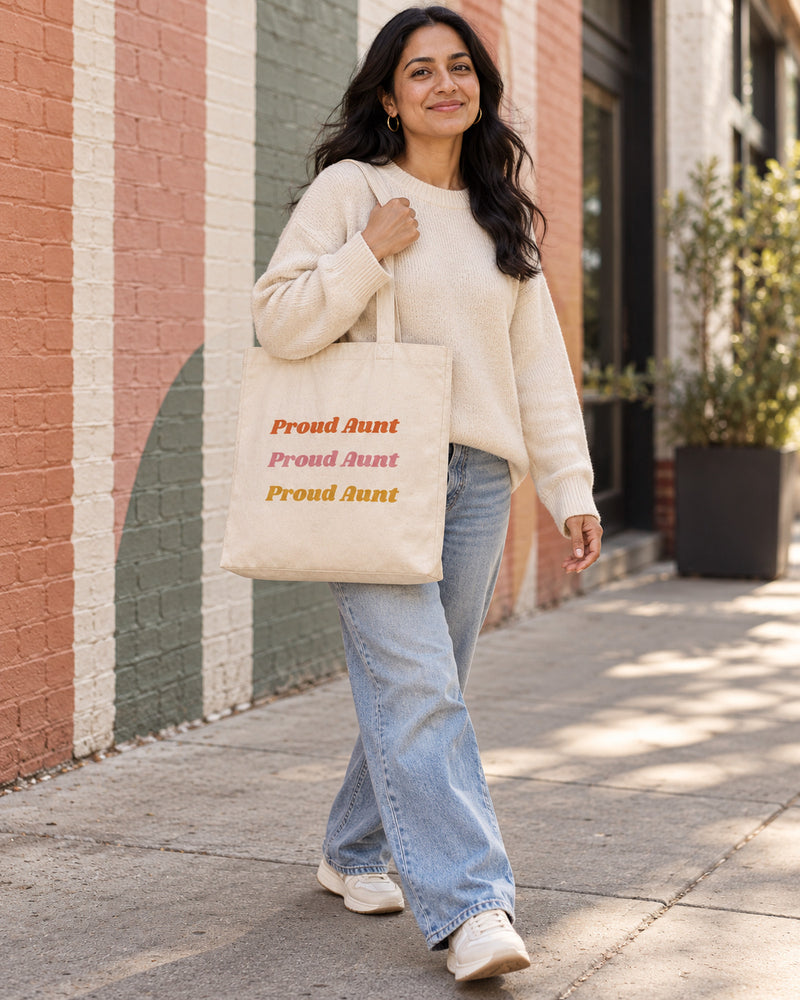 South asian woman carrying a natural canvas tote printed with “Proud Aunt” in orange, rose, marigold. Styled digital image.