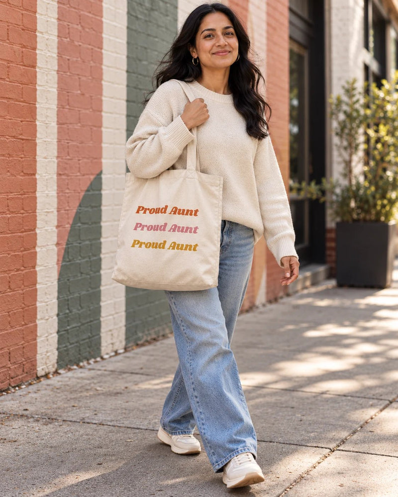 South asian woman carrying a natural canvas tote printed with “Proud Aunt” in orange, rose, marigold. Styled digital image.