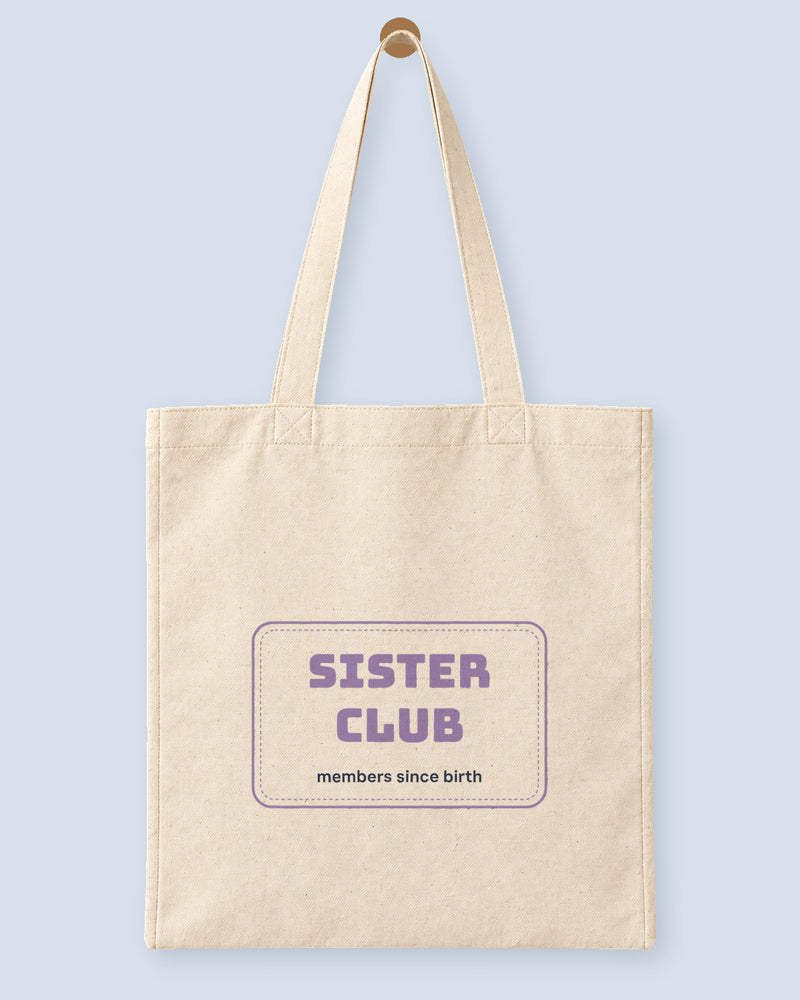 Natural canvas tote printed with “SISTER CLUB”, hanging on a wooden peg.