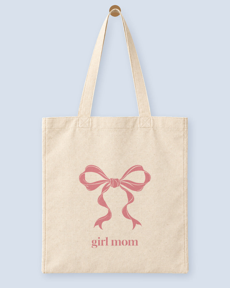 Natural canvas tote printed with “girl mom”, hanging on a wooden peg.