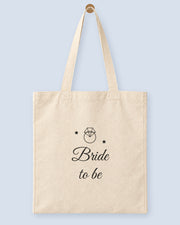 Natural canvas tote printed with “Bride to be”, hanging on a wooden peg.