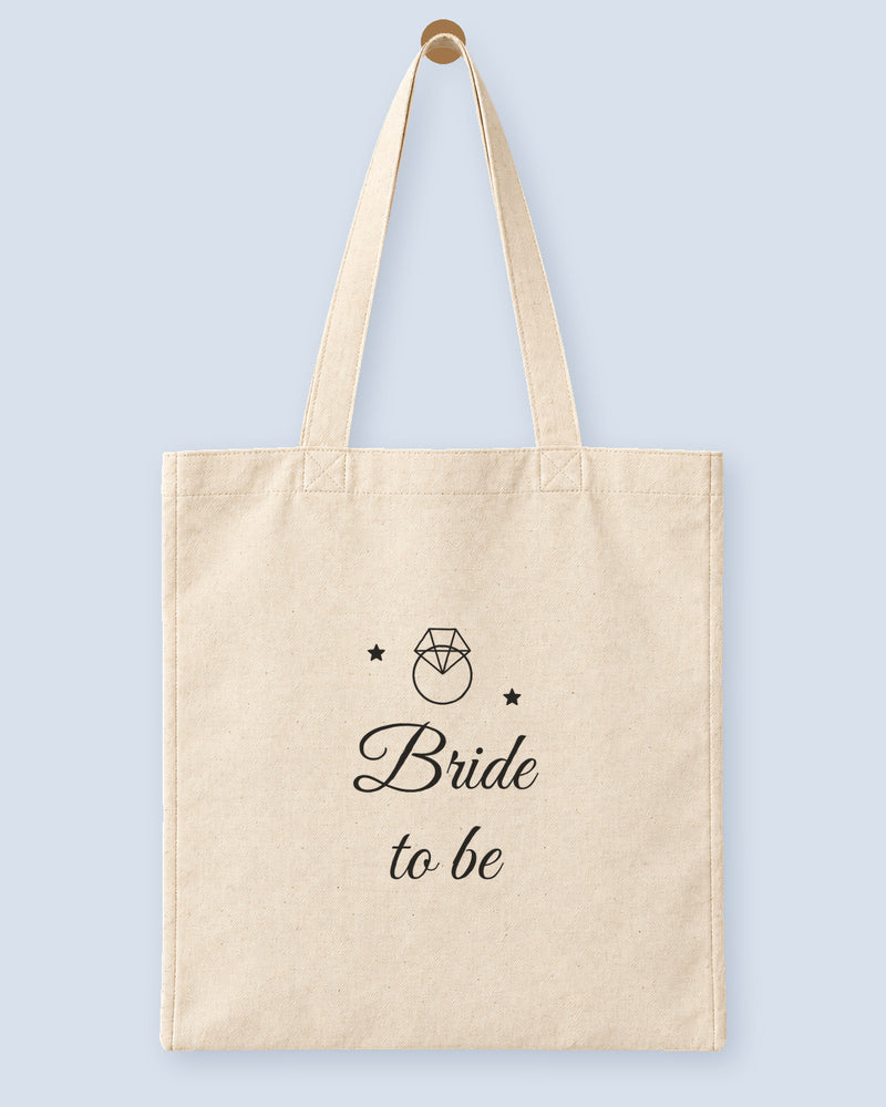 Natural canvas tote printed with “Bride to be”, hanging on a wooden peg.