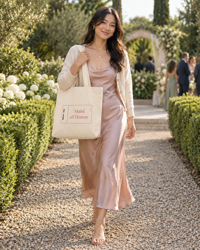 Korean-american woman carrying a natural canvas tote printed with “Maid of Honor” in rose, navy. Styled digital image.