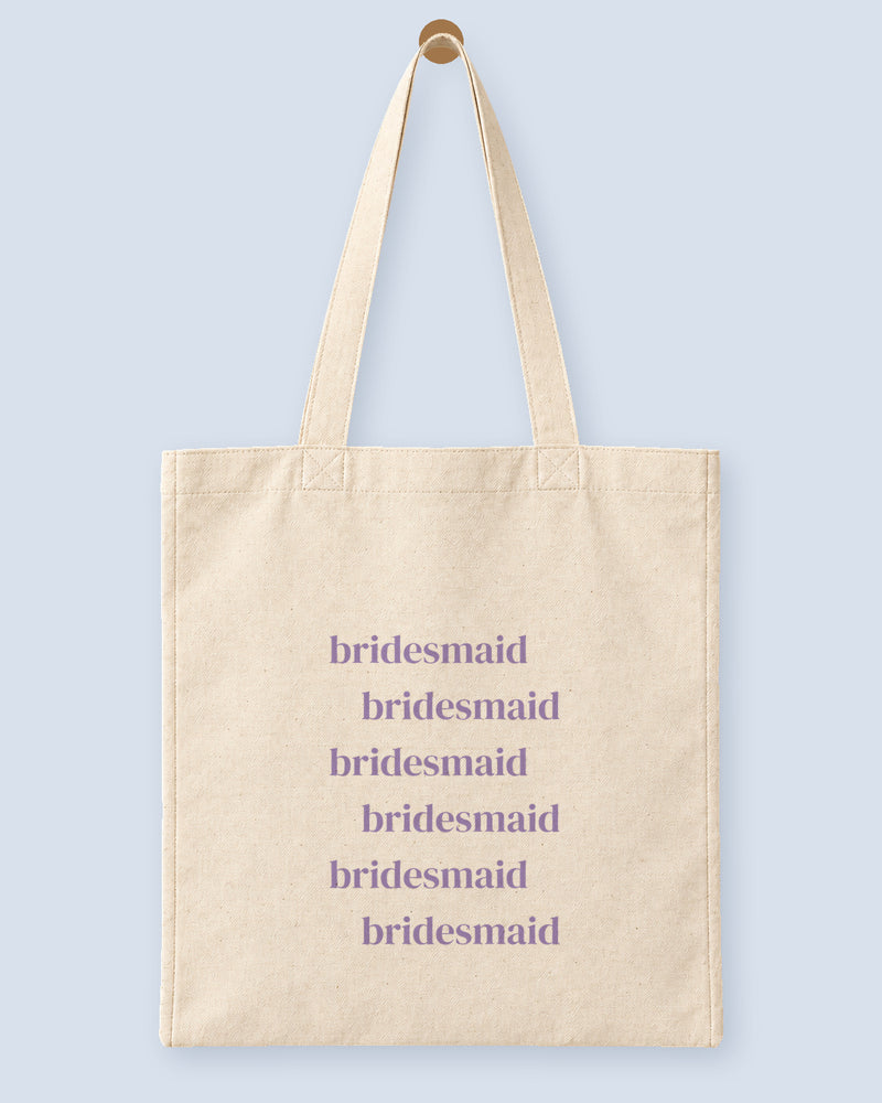 Natural canvas tote printed with “bridesmaid”, hanging on a wooden peg.