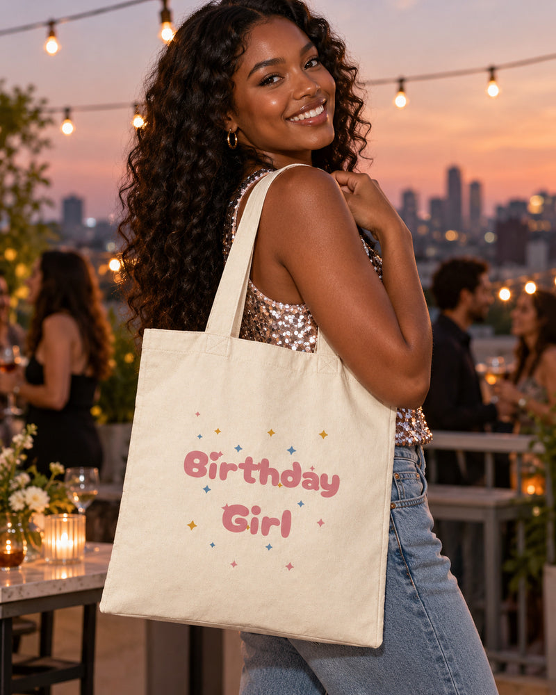Black woman carrying a natural canvas tote printed with “Birthday Girl” in rose, marigold, sky. Styled digital image.