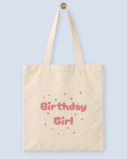 Natural canvas tote printed with “Birthday Girl”, hanging on a wooden peg.