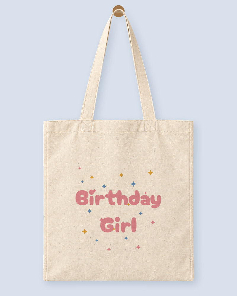 Natural canvas tote printed with “Birthday Girl”, hanging on a wooden peg.