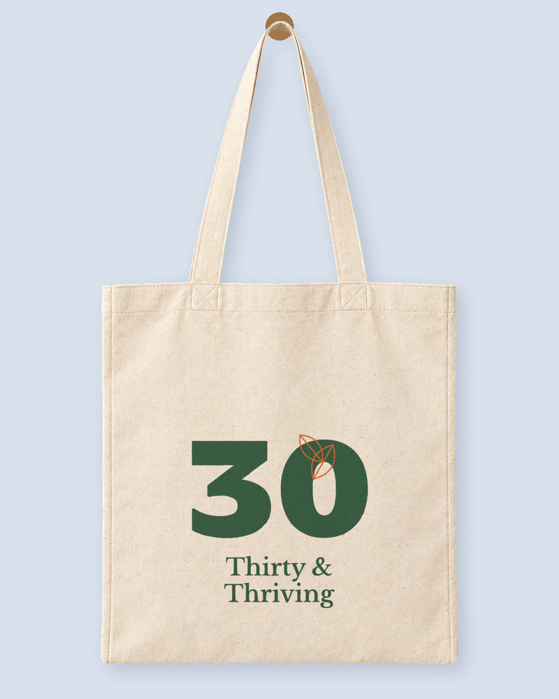 Natural canvas tote printed with “Thirty & Thriving”, hanging on a wooden peg.