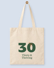 Natural canvas tote printed with “Thirty & Thriving”, hanging on a wooden peg.