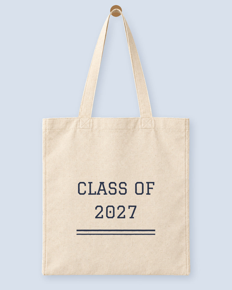 Natural canvas tote printed with “CLASS OF 2027”, hanging on a wooden peg.