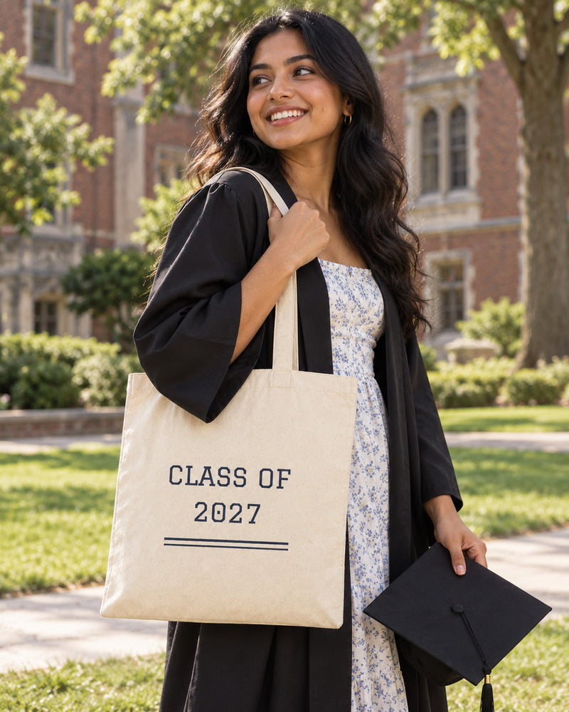 South asian woman carrying a natural canvas tote printed with “CLASS OF 2027” in navy, marigold. Styled digital image.