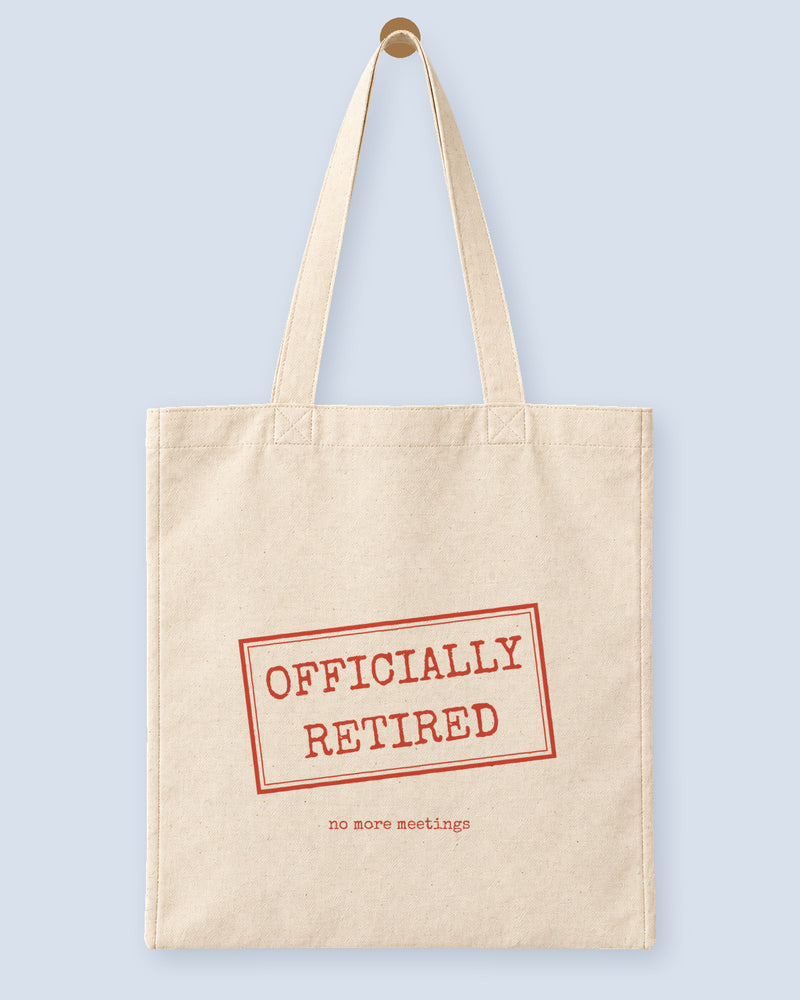 Natural canvas tote printed with “OFFICIALLY RETIRED”, hanging on a wooden peg.