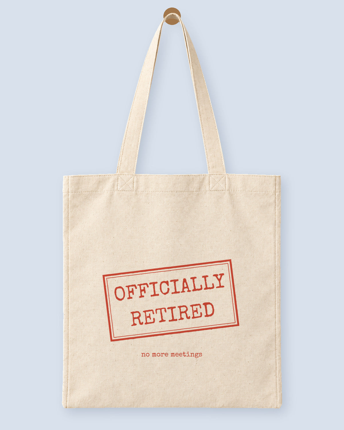 Natural canvas tote printed with “OFFICIALLY RETIRED”, hanging on a wooden peg.