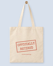 Natural canvas tote printed with “OFFICIALLY RETIRED”, hanging on a wooden peg.