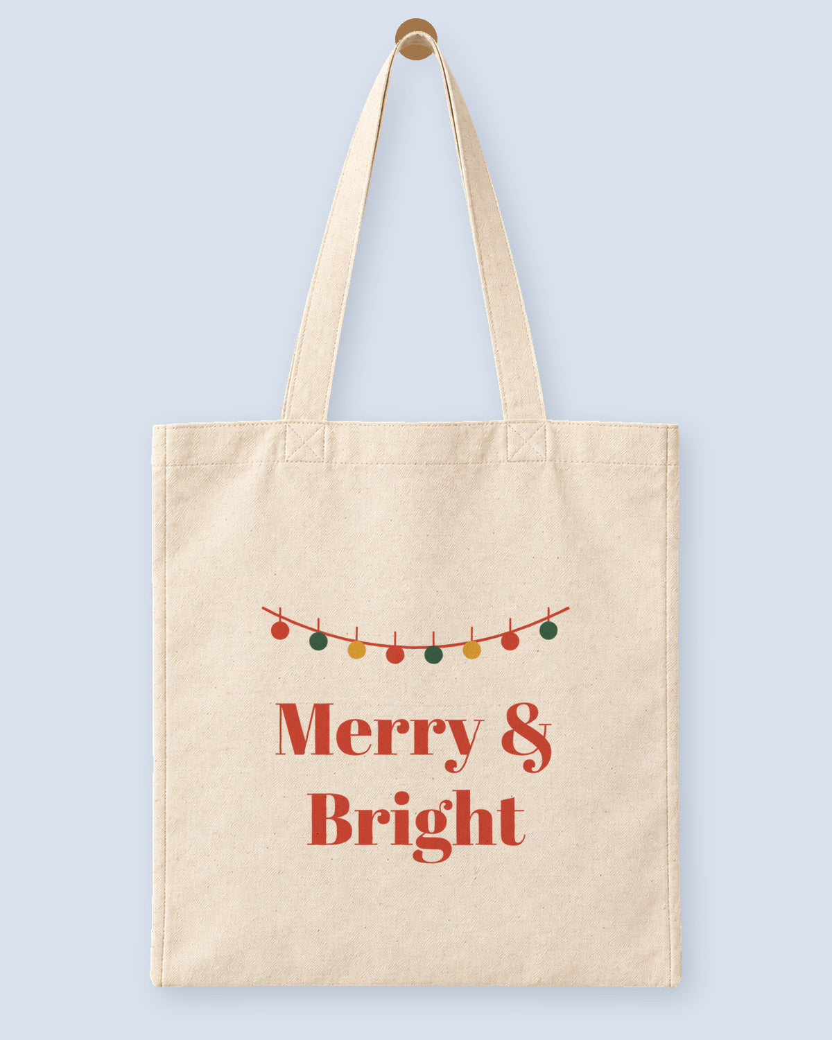 Natural canvas tote printed with “Merry & Bright”, hanging on a wooden peg.