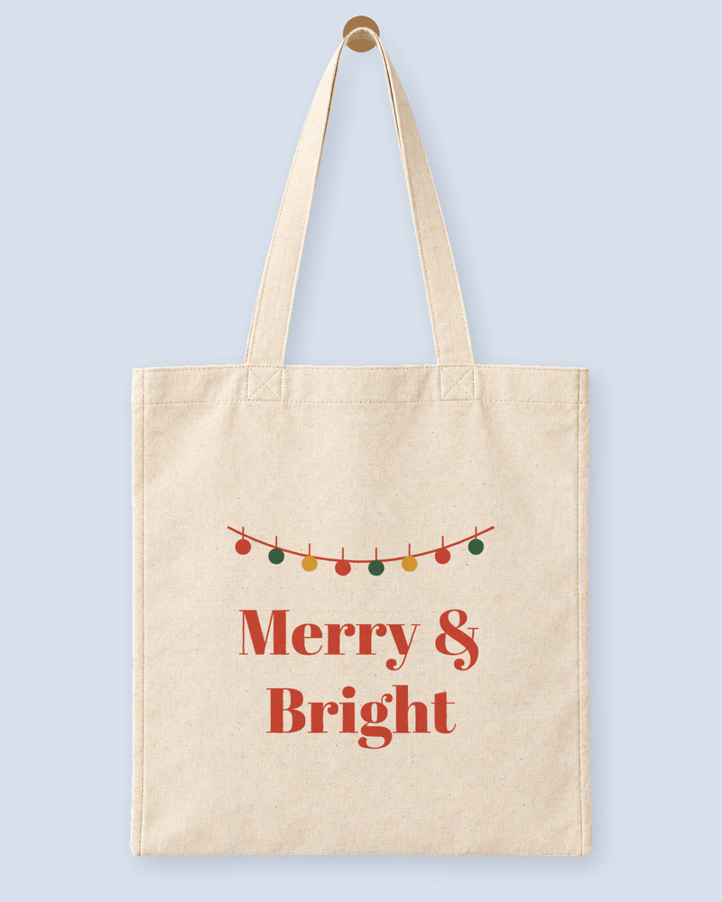 Natural canvas tote printed with “Merry & Bright”, hanging on a wooden peg.
