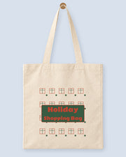 Natural canvas tote printed with “Holiday Shopping Bag”, hanging on a wooden peg.