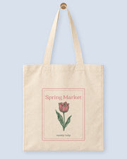 Natural canvas tote printed with “Spring Market”, hanging on a wooden peg.