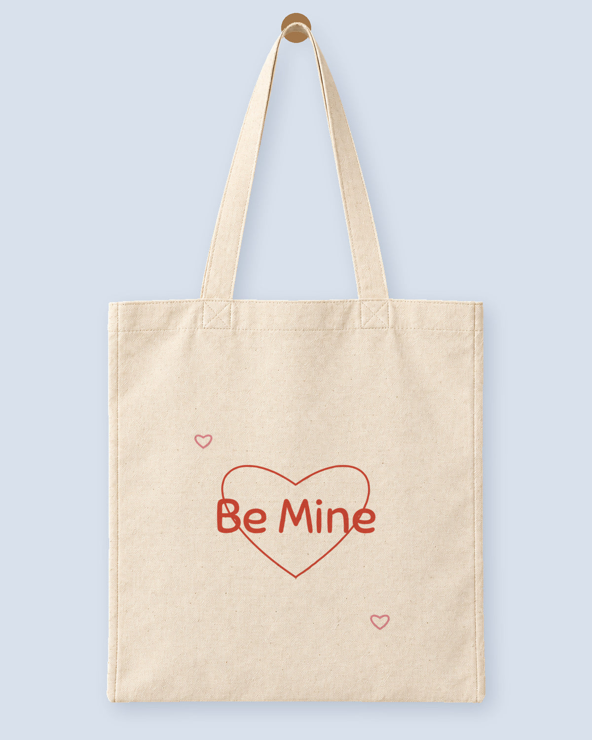 Natural canvas tote printed with “Be Mine”, hanging on a wooden peg.