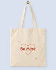 Natural canvas tote printed with “Be Mine”, hanging on a wooden peg.