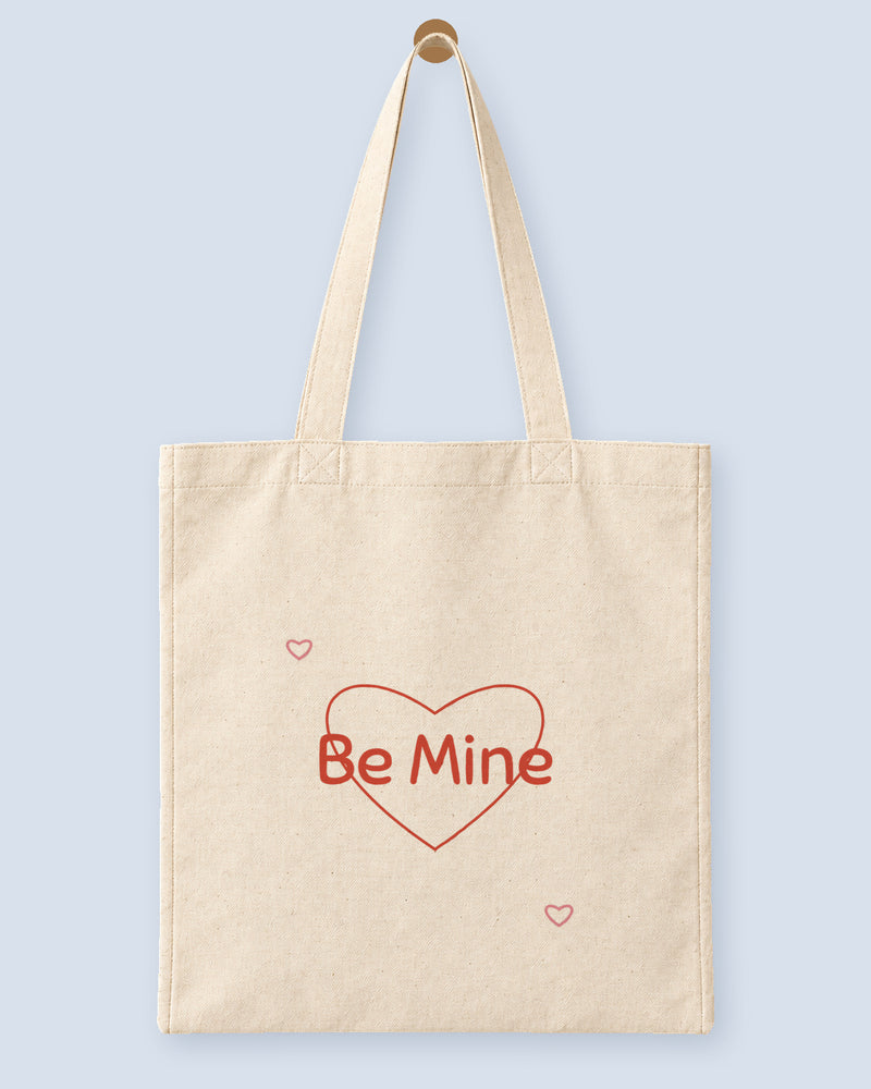 Natural canvas tote printed with “Be Mine”, hanging on a wooden peg.
