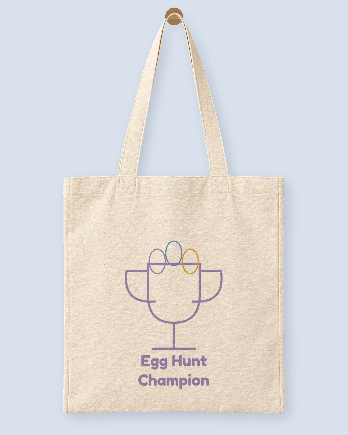 Natural canvas tote printed with “Egg Hunt Champion”, hanging on a wooden peg.