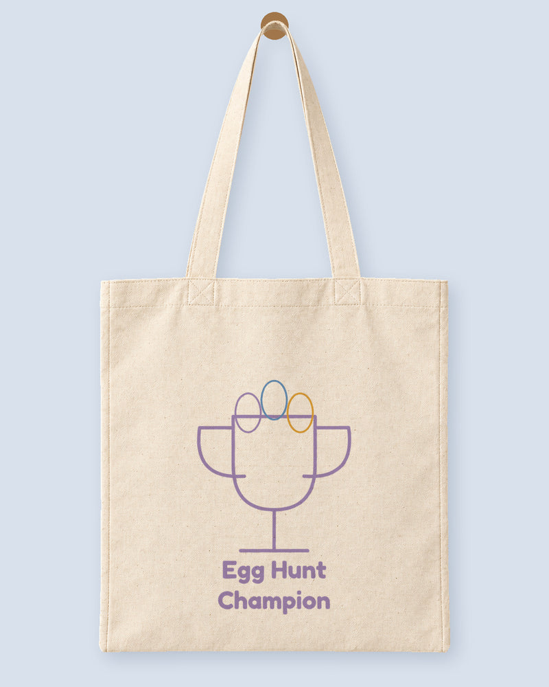 Natural canvas tote printed with “Egg Hunt Champion”, hanging on a wooden peg.