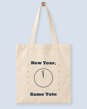 Natural canvas tote printed with “New Year, Same Tote”, hanging on a wooden peg.