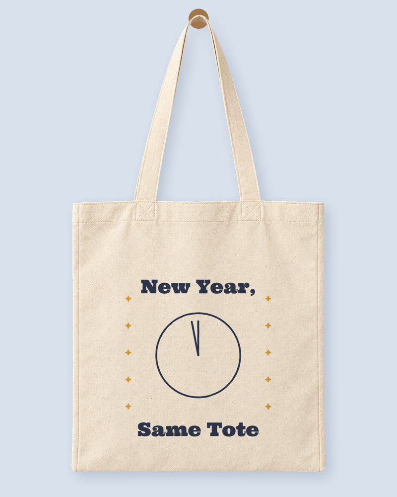 Natural canvas tote printed with “New Year, Same Tote”, hanging on a wooden peg.