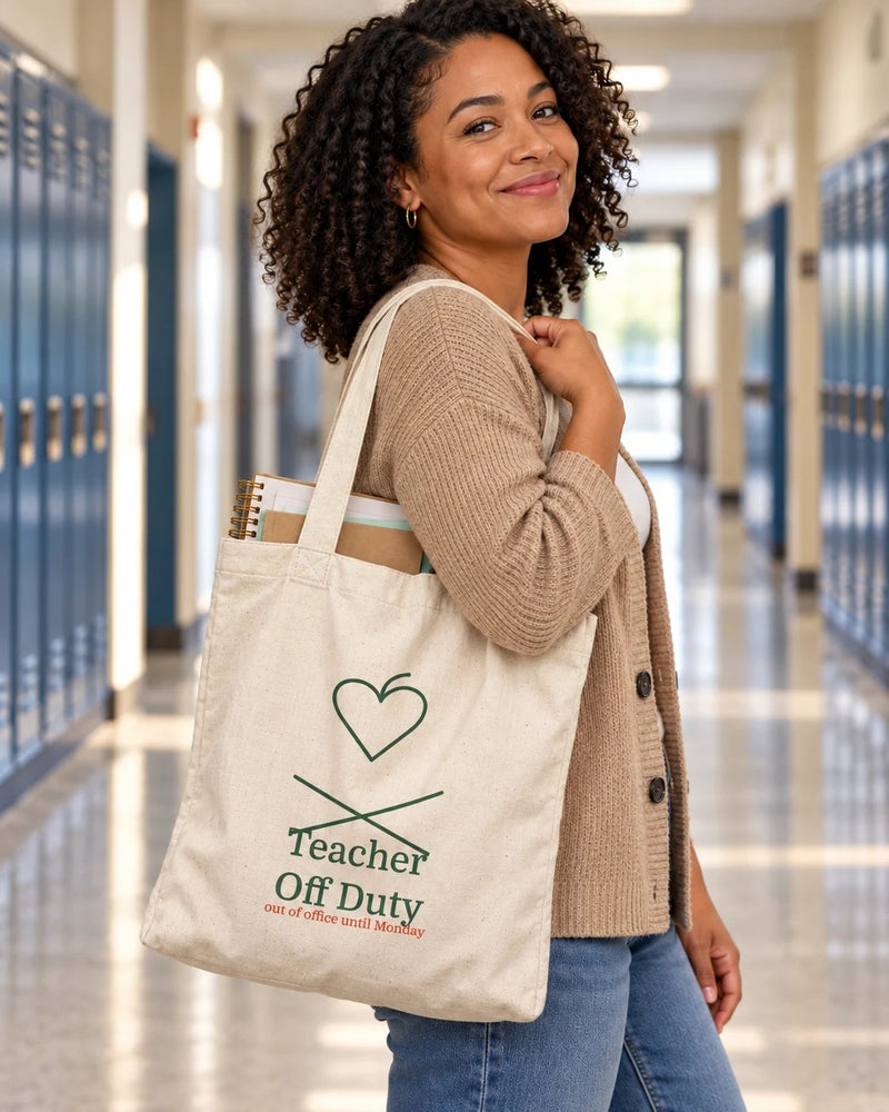 Black woman carrying a natural canvas tote printed with “Teacher Off Duty” in forest, red. Styled digital image.