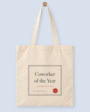 Natural canvas tote printed with “Coworker of the Year”, hanging on a wooden peg.