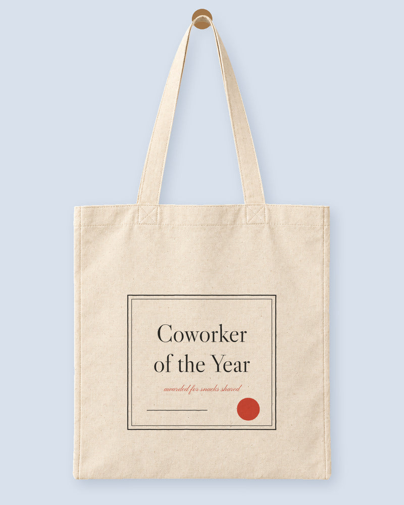 Natural canvas tote printed with “Coworker of the Year”, hanging on a wooden peg.