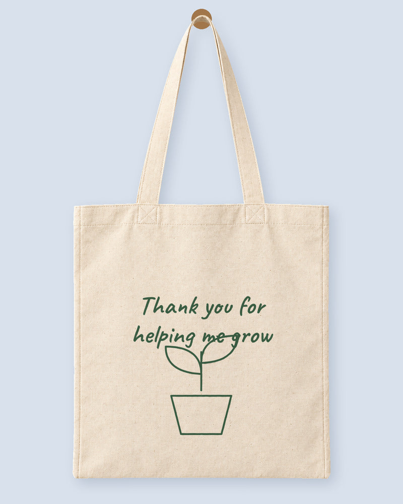 Natural canvas tote printed with “Thank you for helping me grow”, hanging on a wooden peg.