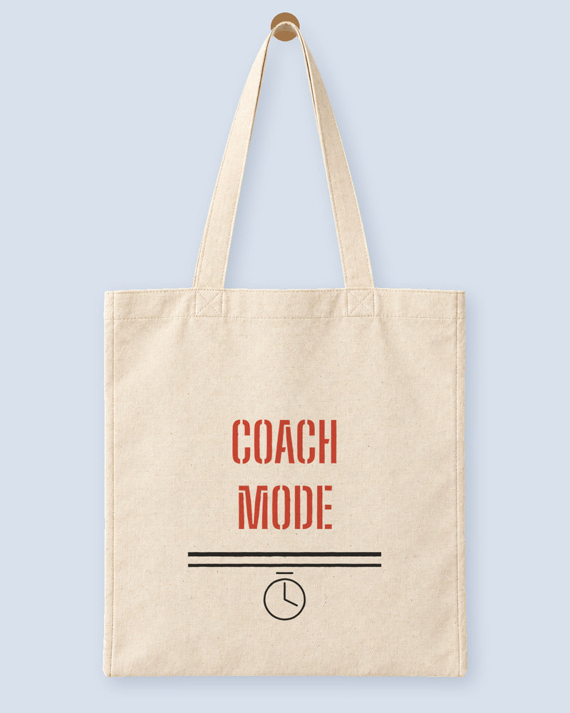 Natural canvas tote printed with “COACH MODE”, hanging on a wooden peg.