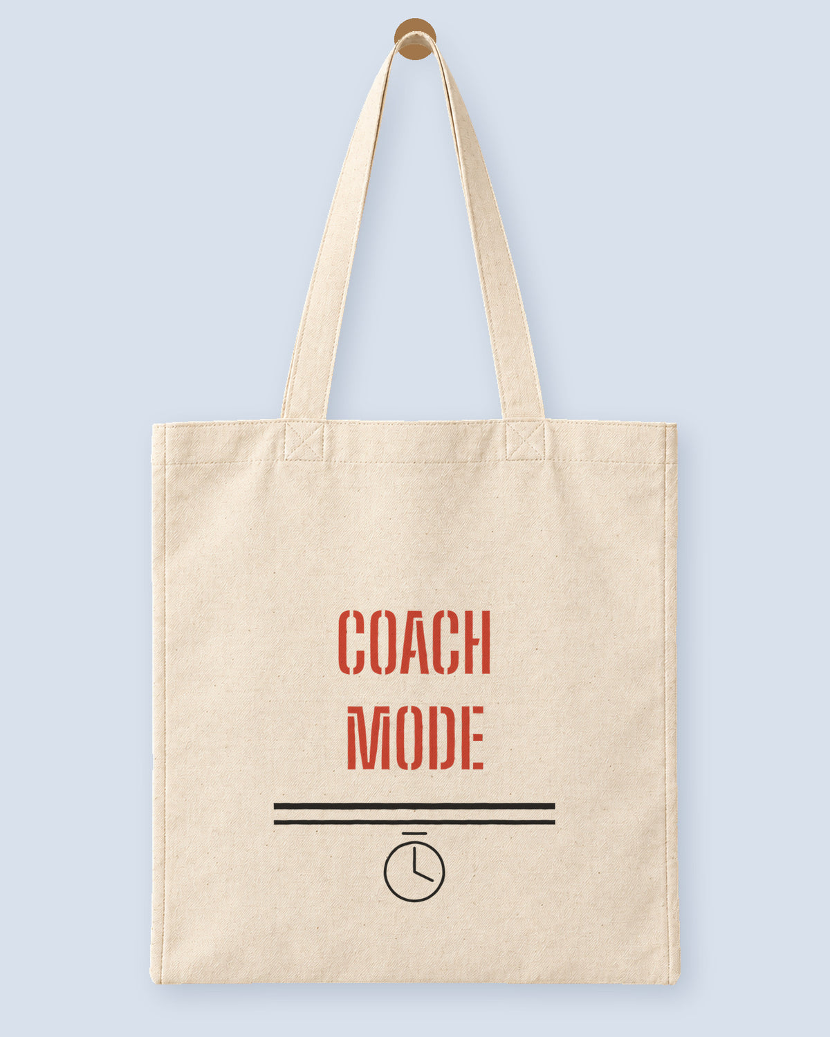 Natural canvas tote printed with “COACH MODE”, hanging on a wooden peg.