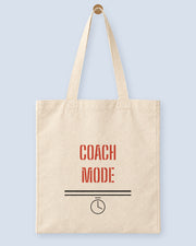 Natural canvas tote printed with “COACH MODE”, hanging on a wooden peg.