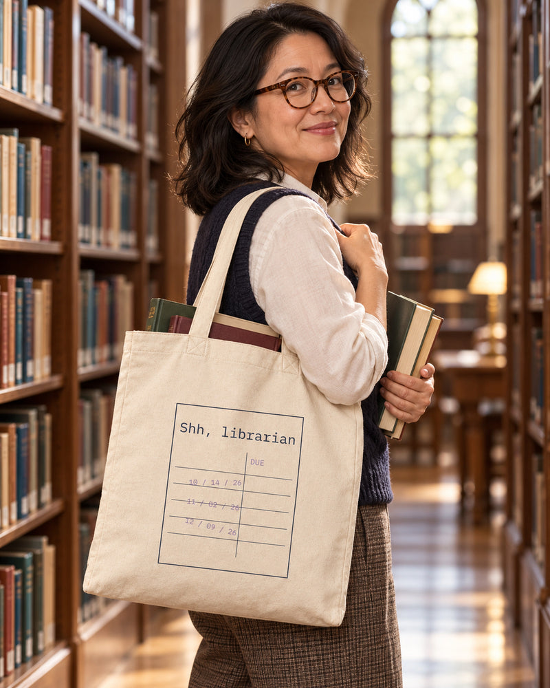 Chinese-american woman carrying a natural canvas tote printed with “Shh, librarian” in navy, lilac. Styled digital image.