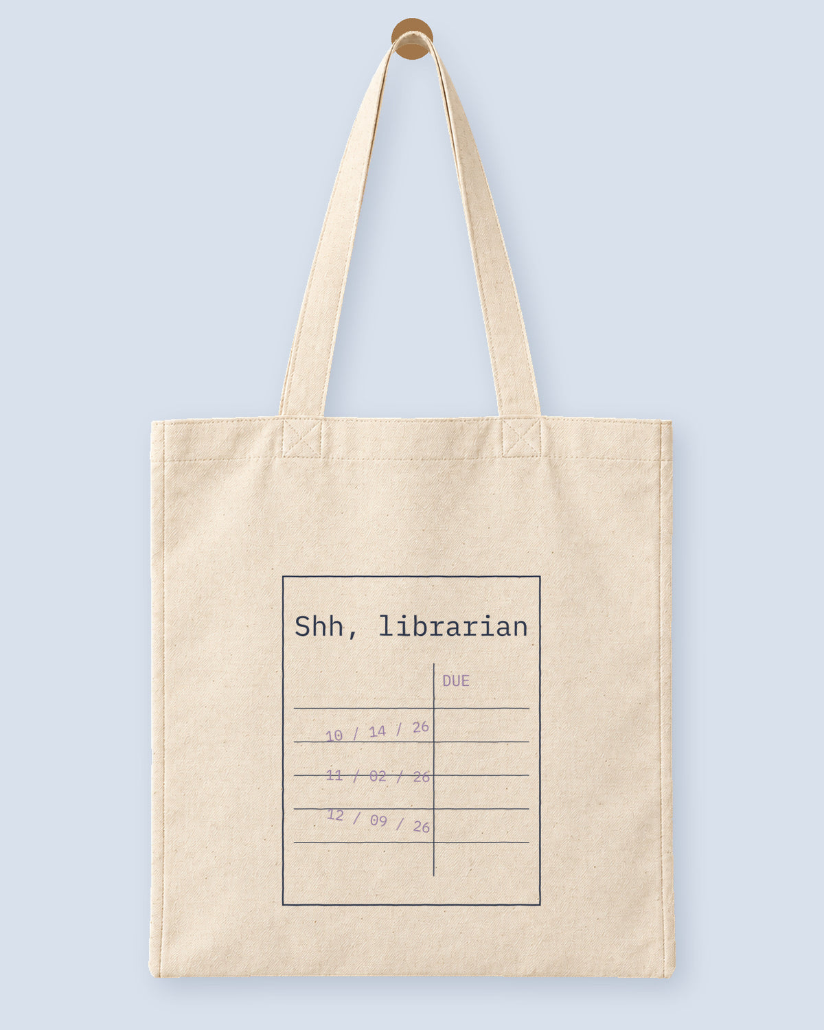 Natural canvas tote printed with “Shh, librarian”, hanging on a wooden peg.
