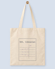 Natural canvas tote printed with “Shh, librarian”, hanging on a wooden peg.